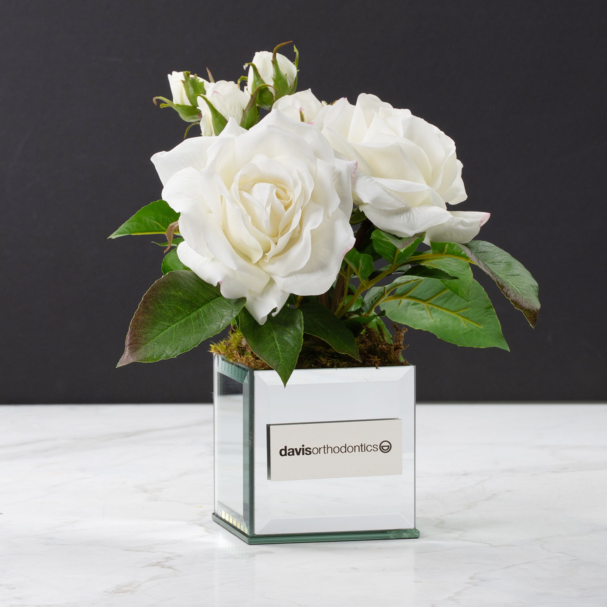 The Audrey faux floral arrangement of white roses with green leaves in a clear square glass vase with a white label reading 'davis orthodontics', set on a white marble surface against a dark gray background.