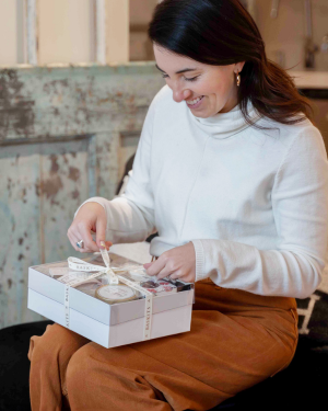 A woman in a white turtleneck sweater and brown skirt smiles while opening a white gift box tied with a ribbon.